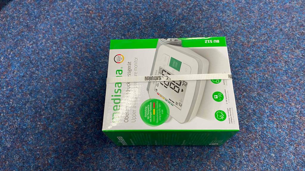 Im Test: Ein verpacktes Medisana-Blutdruckmessgerät liegt horizontal auf einem Teppichboden.