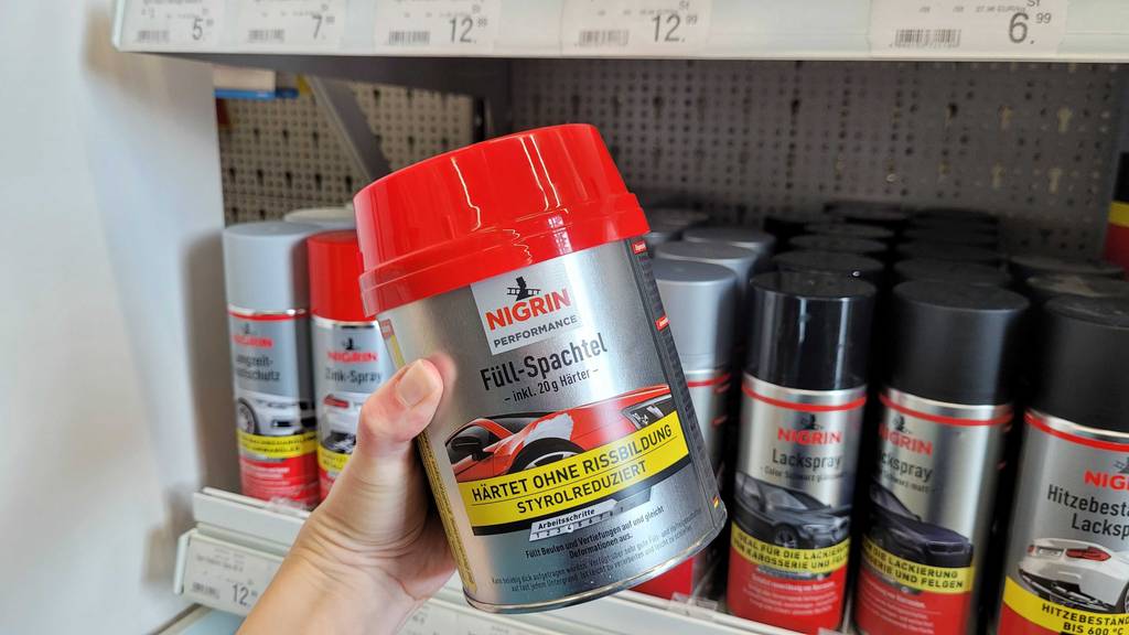Spachtelmasse (Auto) getestet: Eine Hand hält eine Dose Spachtelmasse (Auto) vor ein Regal.