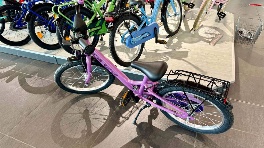 Das getestete Mädchenfahrrad 18 Zoll im pinkfarbenen Design steht vor einem Ausstellungspodest mit Fahrrädern.