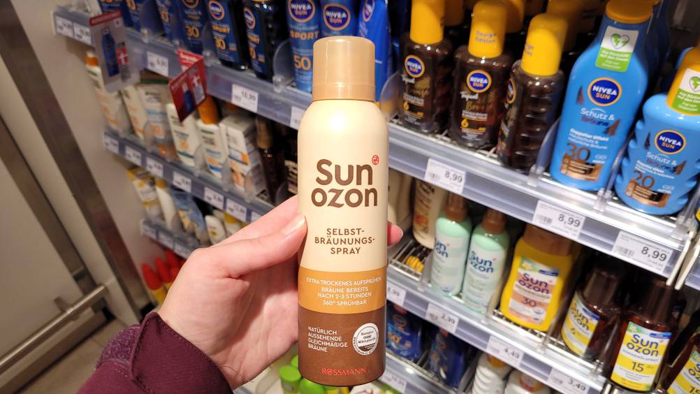 Bräunungsspray getestet: Eine Hand hält ein Bräunungsspray vor ein Regal mit Produkten.