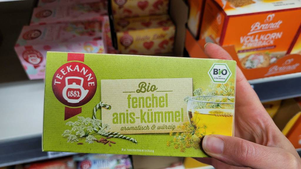 Im Test: Kümmeltee ine einer Teekanne-Verpackung in einer Hand vor einem Regal mit weiteren Teesorten.