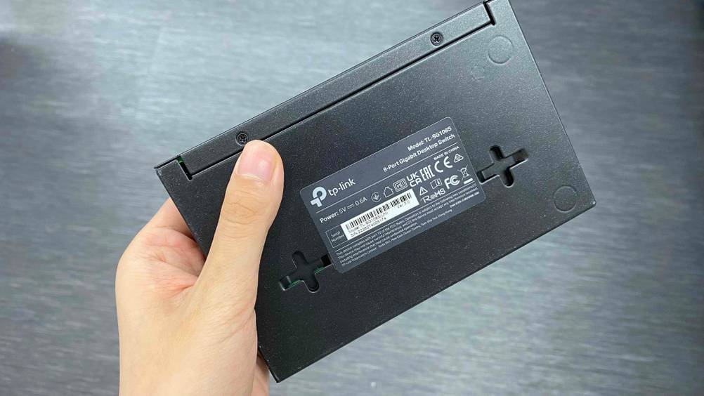 Eine Hand hält die Rückseite mit Informationen des 8-Port-Switches vor eine graue Fläche.