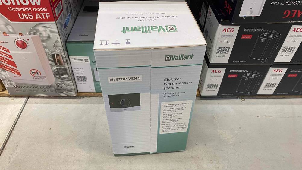 Ein getesteter Vaillant Warmwasserspeicher in Verpackung steht auf einem hellen Boden vor anderen Produkten.