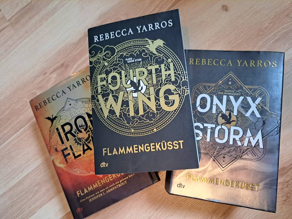 3 Bücher liegen als Pyramidenstapel auf einem Holzboden. Die drei Bücher sind die 3 Bänder der Fouth Wing-Reihe von Rebecca Yarros. Das obere Buch ist ein Taschenbuch, die anderen beiden sind mit Harteinband.