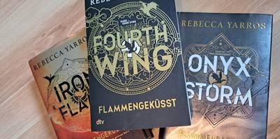 3 Bücher liegen als Pyramidenstapel auf einem Holzboden. Die drei Bücher sind die 3 Bänder der Fouth Wing-Reihe von Rebecca Yarros. Das obere Buch ist ein Taschenbuch, die anderen beiden sind mit Harteinband.
