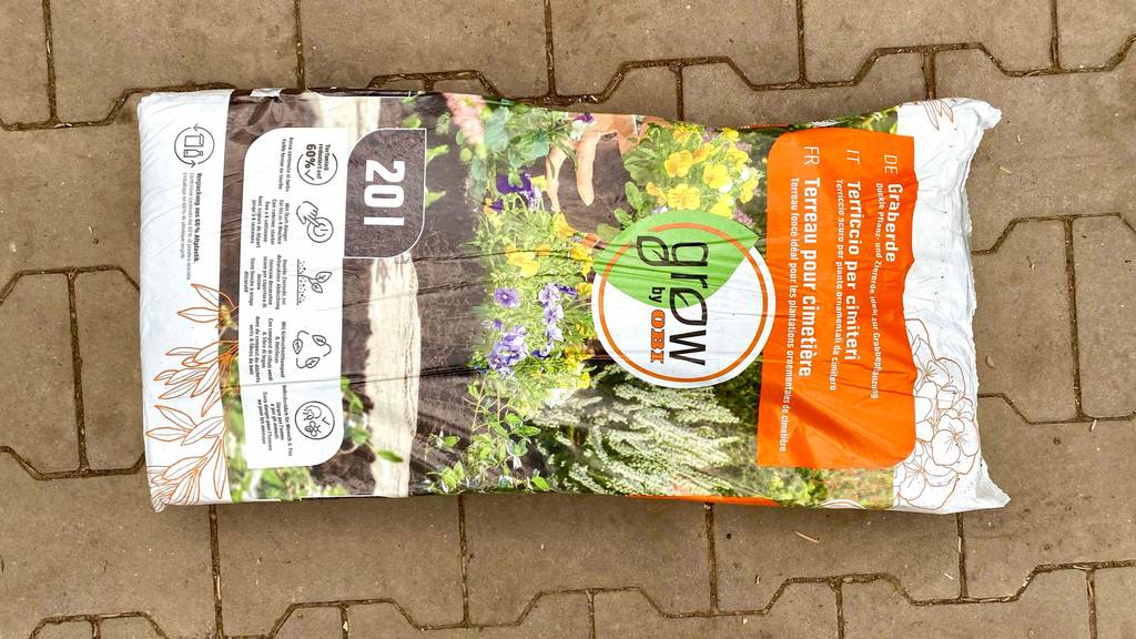 Ein 20-Liter-Kunststoffsack inklusive Graberde im Test von Grow by Obi liegt auf rot-braunen Pflastersteinen.