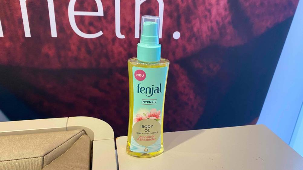 Eine Flasche des getesteten Körperöls auf einer Fläche vor einem Werbeplakat.