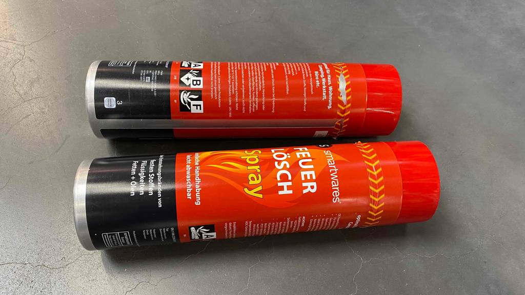 Feuerlöschspray getestet: Zwei Dosen liegen horizontal nebeneinander auf einer grauen Fläche