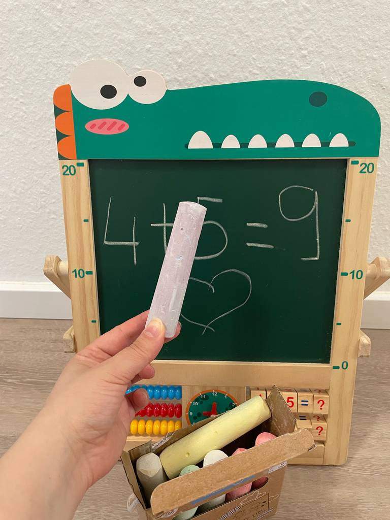 Kreidestifte-Test: Eine Hand hält eine Malkreide vor einer bemalten grünen Tafel.