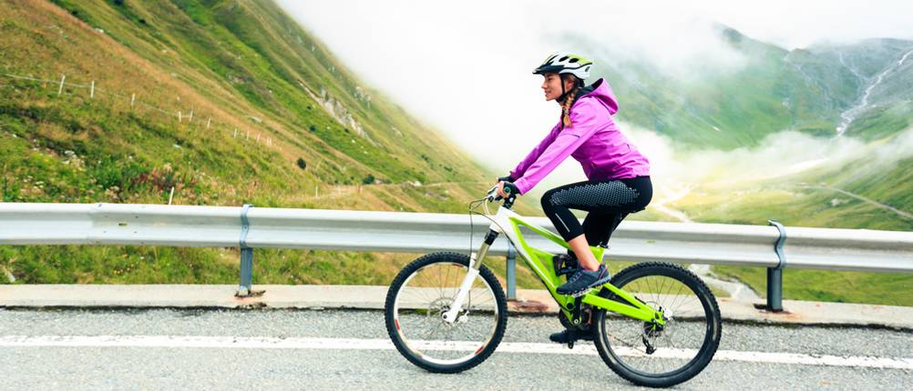 3/4-Radhose-Damen-Test: eine Frau in 3/4-Radhose fährt auf einem Mountainbike auf einer Straße; im Hintergrund sind Berge und Nebelschwaden zu sehen.