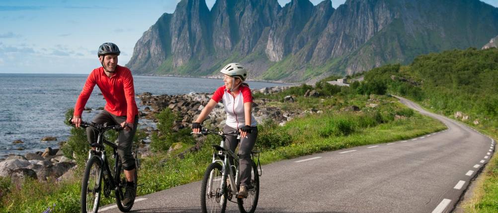 3/4-Radhosen-Herren-Test: Ein Mann in 3/4-Hose radelt neben einer Frau an einem See oder Meer entlang auf einer Straße. Im Hintergrund sind Berge zu sehen.