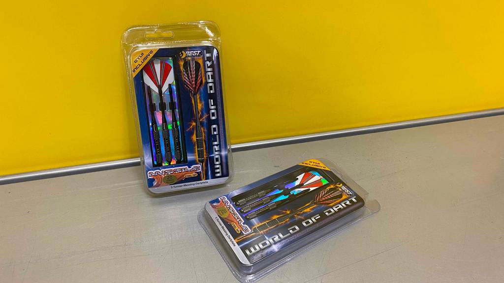 Zwei Packungen getestete Steeldarts, eine lehnt an einer Wand eine liegt auf dem grauen Boden.