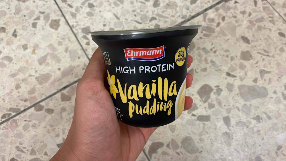 Protein Pudding im Test: Eine Hand hält einen Becher Pudding über einen Fließenboden