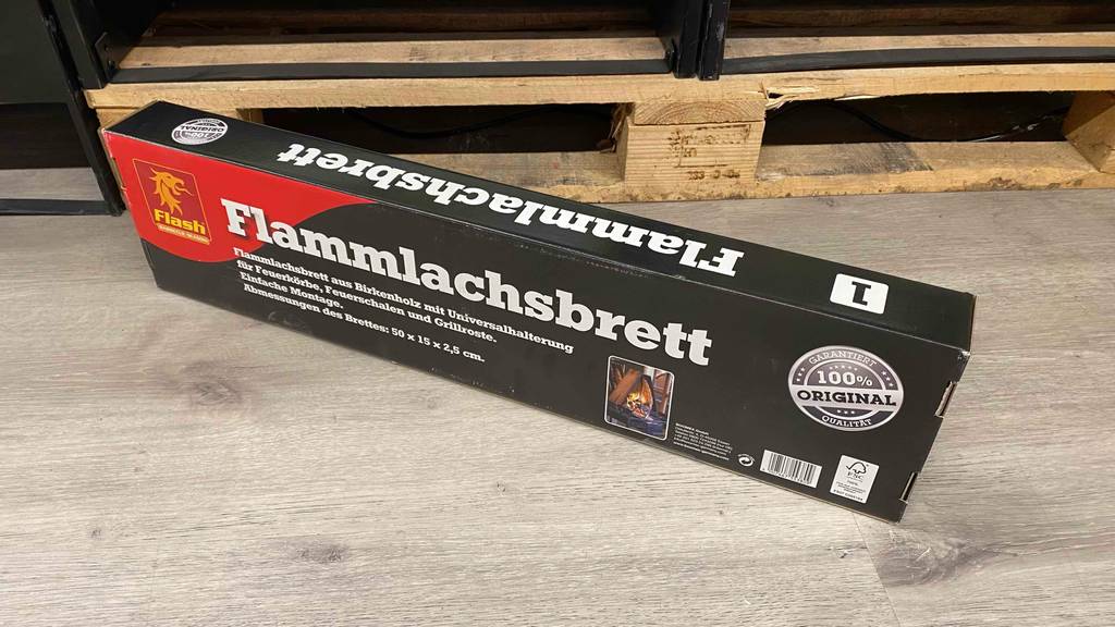 Flammlachsbrett getestet: Ein verpacktes Flammlachsbrett liegt vor einer Holzpalette auf einem grauen Boden.