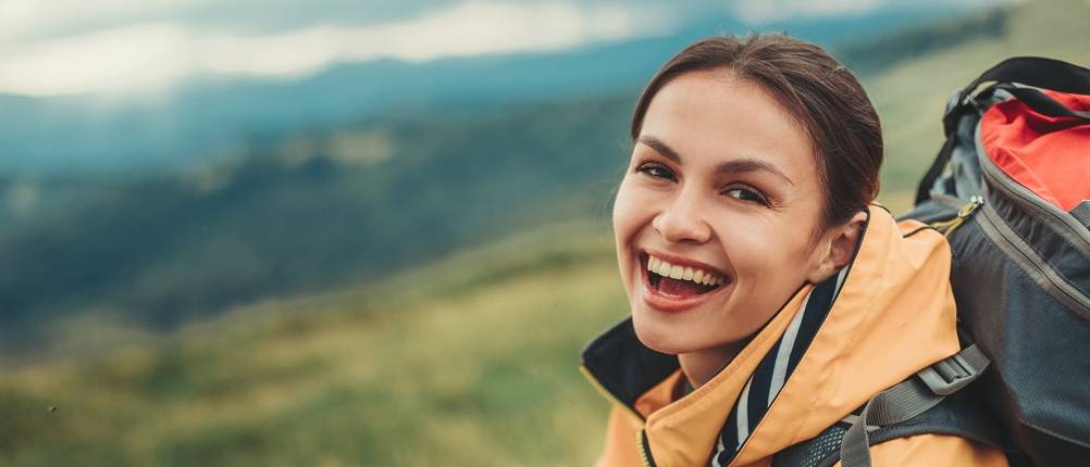 3-in-1-Damen-Jacken-Test: Eine lachende Frau im Gebirge mit einer Wanderjacke in Orange und einem Rucksack.