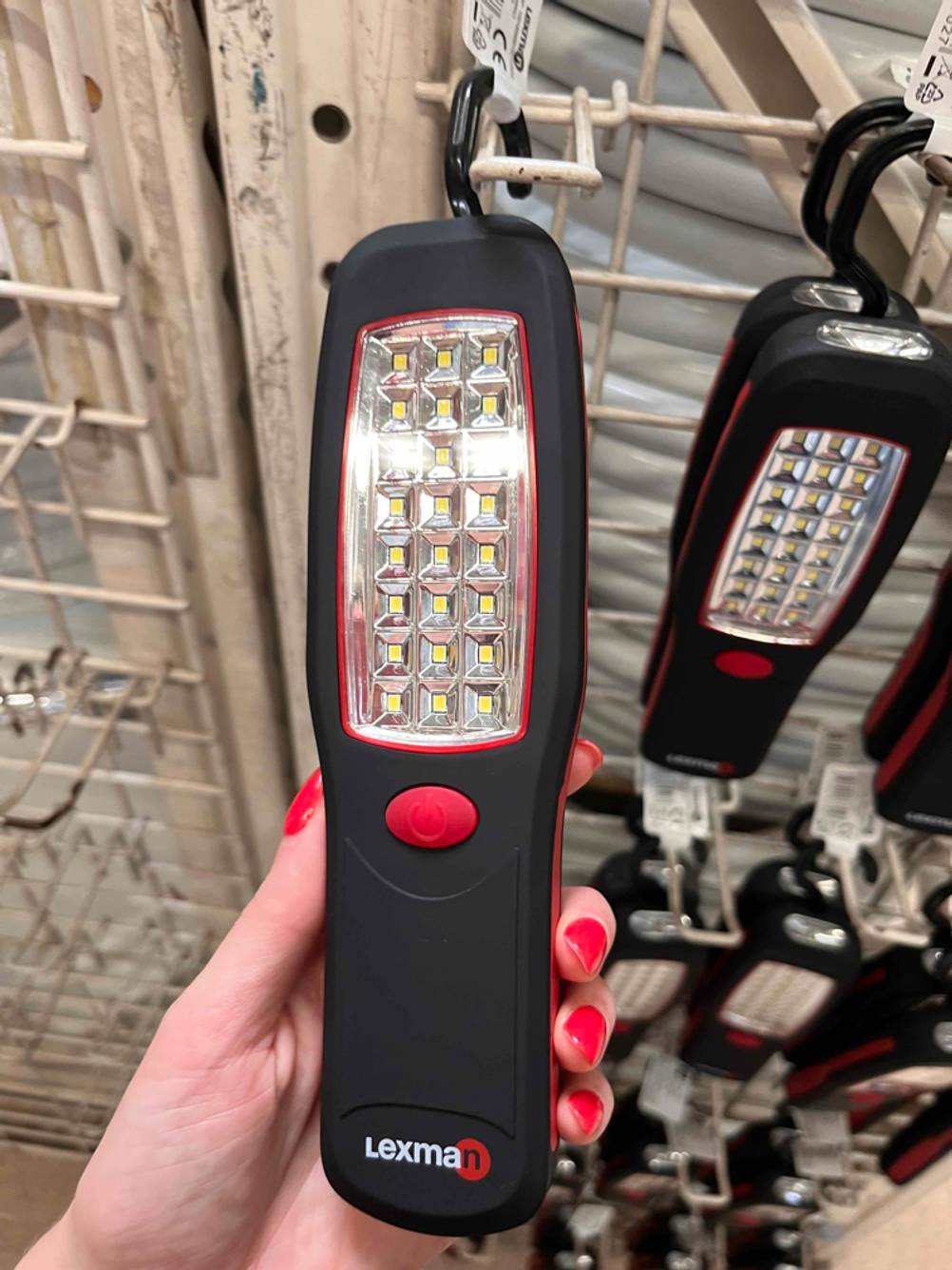 LED-Strahler-Test: Hand hält schwarzen LED-Strahler von Lexman mit rotem Einschaltknopf vor Kaufregal. Blick auf die Vorderseite.