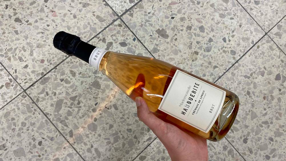 Crémant getestet: Eine Hand hält eine Flasche Crémant vor einen hellen Fließenboden