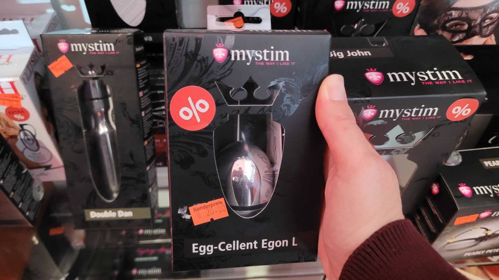 Eine Hand hält ein getestetes Vibro-Ei in seiner Origialverpackung vor ein Regal.