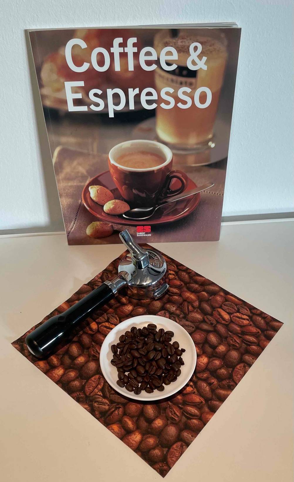 Ein Buch lehnt an der Wand, davor eine Serviette auf der ein Teller mit getesteten Espressobohnen liegt und ein Siebträger.