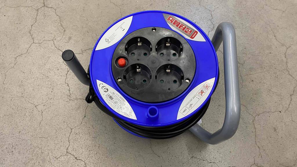 Blaue getestete Kabeltrommel liegt auf grauem Boden und gibt Sicht frei auf vier Steckdosen und einen silberfarbenen Griff.