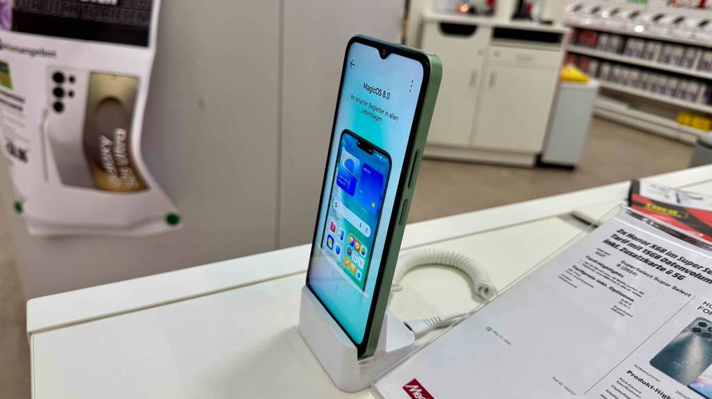 Das getestete Smartphone unter 100 Euro von der Seite betrachtet, mit Fokus auf das eingeschaltete Display, steht in einem Verkaufsbereich.