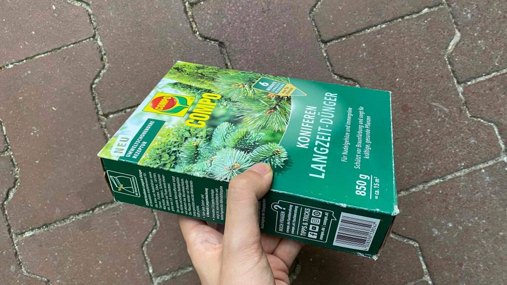 Koniferendünger im Test: Eine Hand hält eine Packung vor einen dunklen Hintergrund