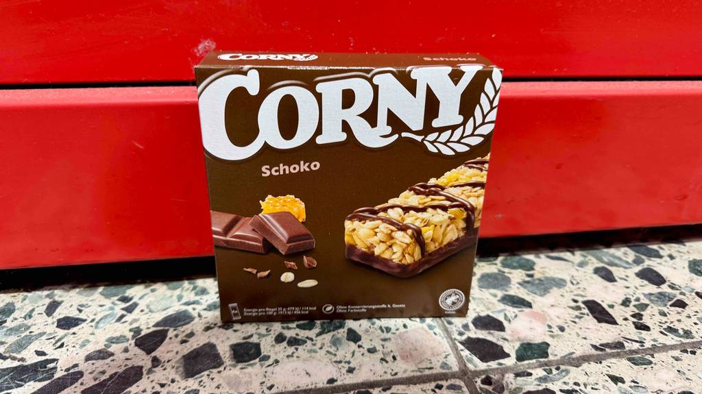 Eine Packung der getesteten Corny-Riegel in der Geschmacksrichtung Schoko steht vor einem roten Hintergrund.