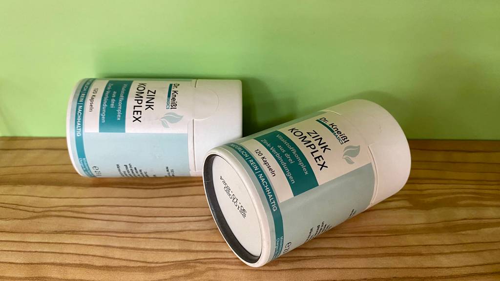 Zink im Test: Zwei Dosen Zink-Komplex liegend auf einer Holzfläche vor grüner Wand.