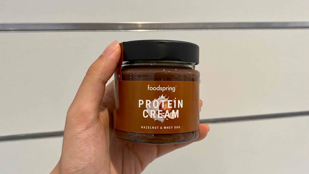 Protein Creme getestet: Eine Hand hält ein Glas Protein Creme vor eine helle Oberfläche