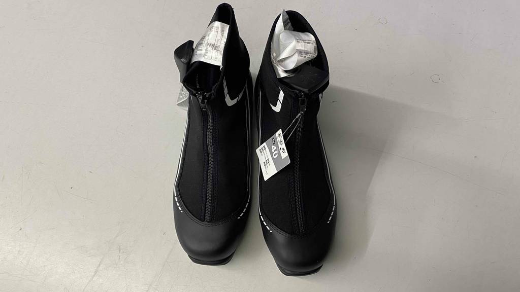 Langlaufschuh getestet: schwarze Langlaufschuhe mit einem Reißverschluss stehen auf einer weißen Fläche mit Eitkett darauf.