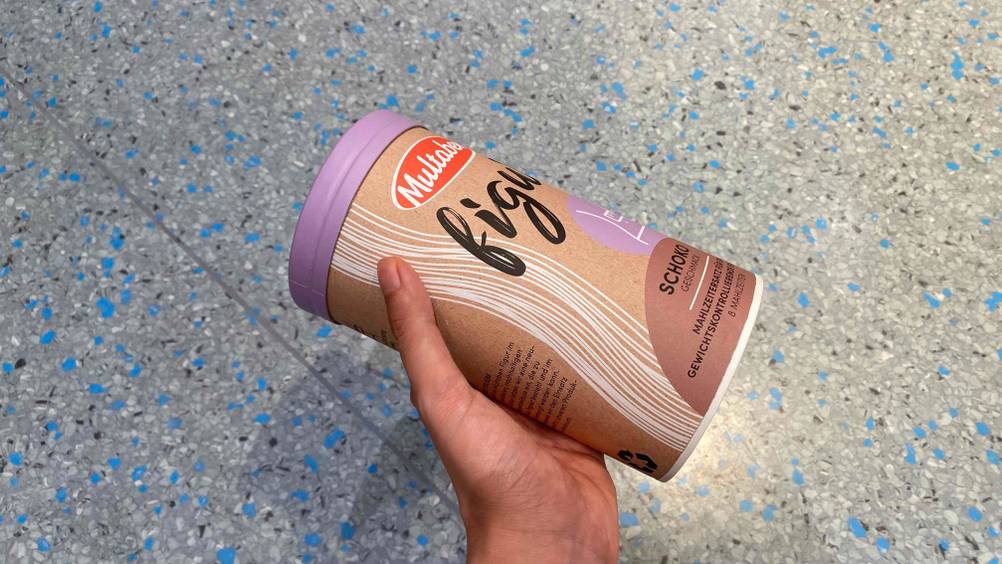 Im Test: Eine Hand hält eine Packung Eiweißpulver vor einen Fließenboden
