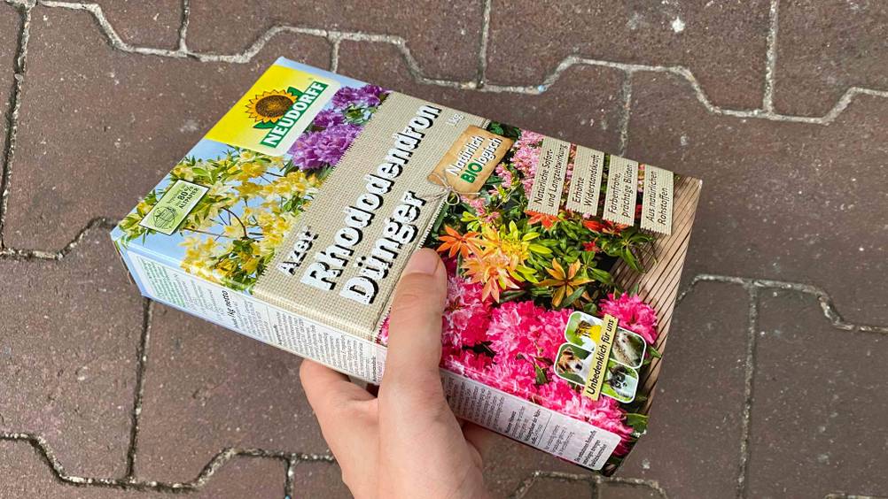 Rhododendron-Dünger im Test: Eine Hand hält eine Packung vor einen Steinboden