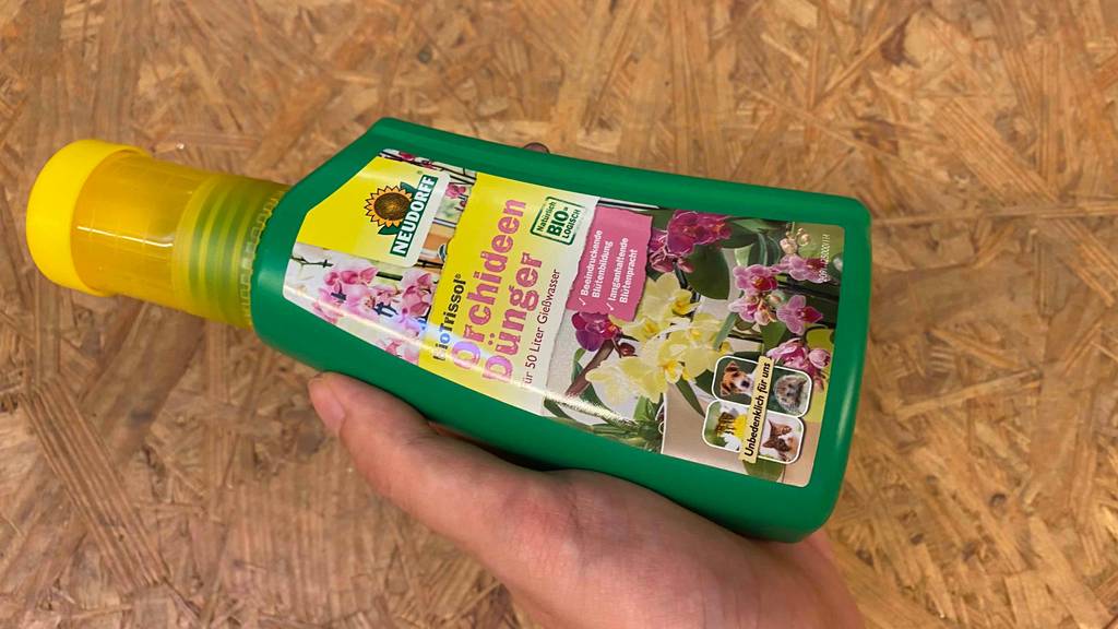 Orchideendünger im Test: Eine Hand hält eine Flasche Dünger vor eine Holzfläche