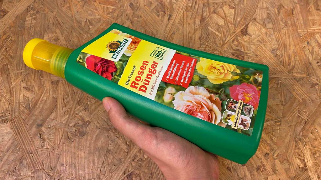 Rosendünger im Test: Eine Hand hält eine Flasche Dünger vor eine Holzfläche