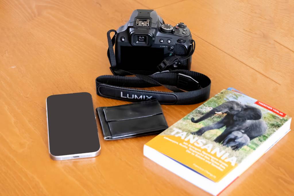 Die getestete Reisezoom-Kamera liegt neben einem Buch, Smartphone und einer Geldtasche.