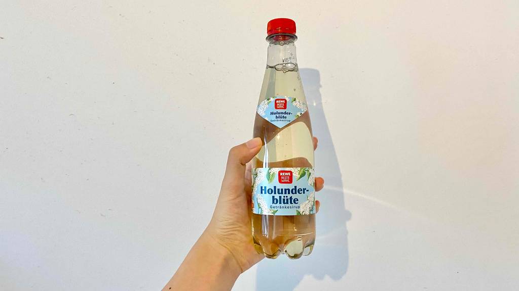 Getesteter Holunderblütensirup in der Hand vor weißer Wand.
