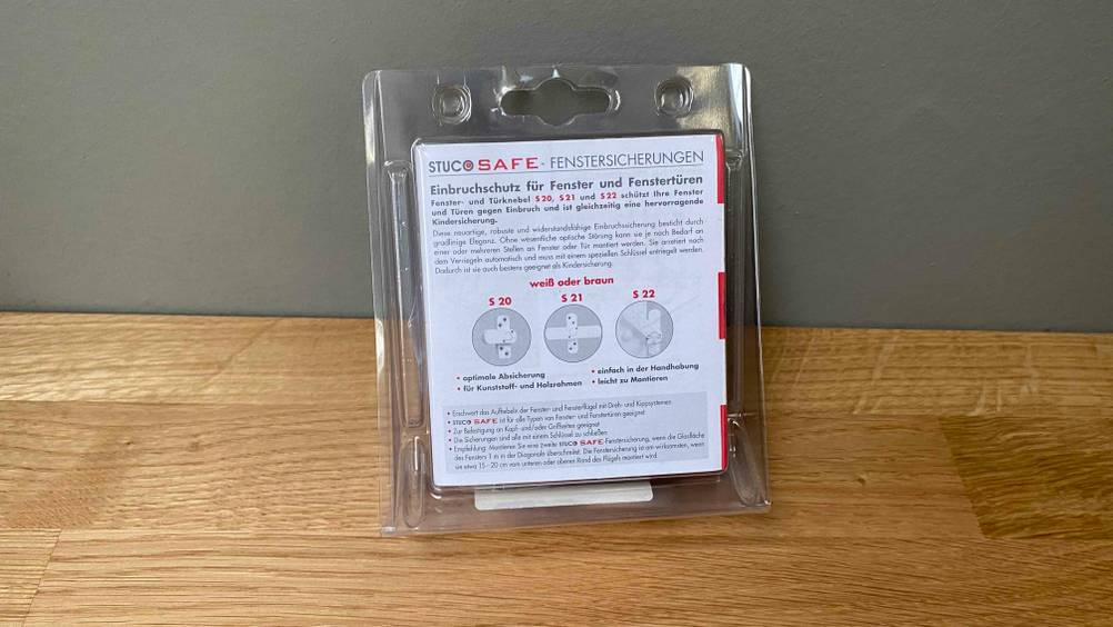 Im Test: Eine Fenstersicherung lehnt in ihrer Verpackung an eine graue Wand.