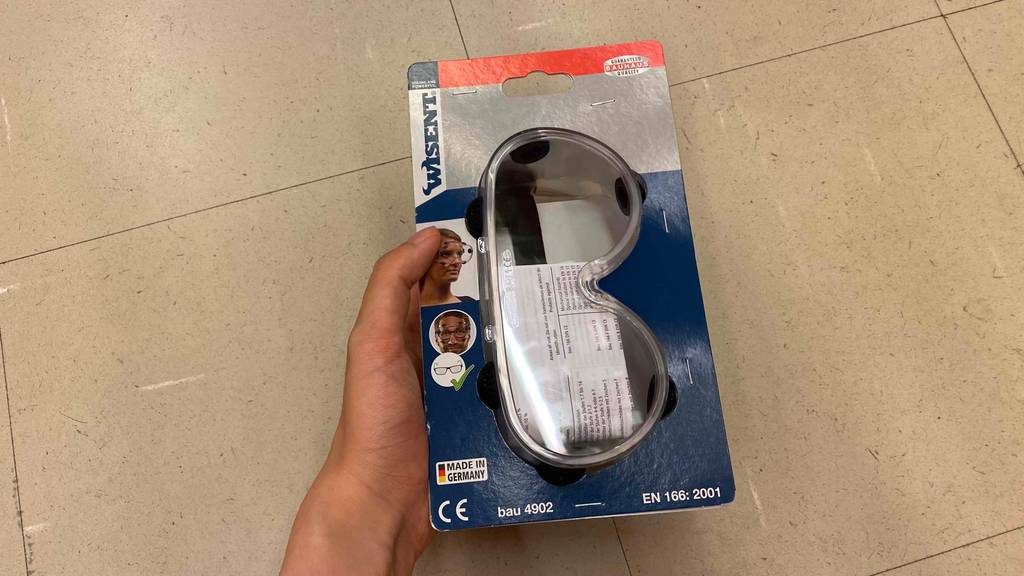 Schutzbrille getestet: Eine Hand hält eine Brille in Originalverpackung vor einen Fließenboden