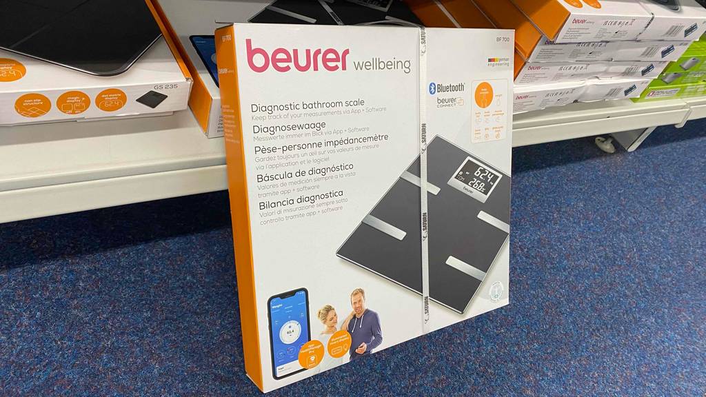 Im Test: Eine verpackte Körperfettwaage mit App lehnt an einem Regalbrett.