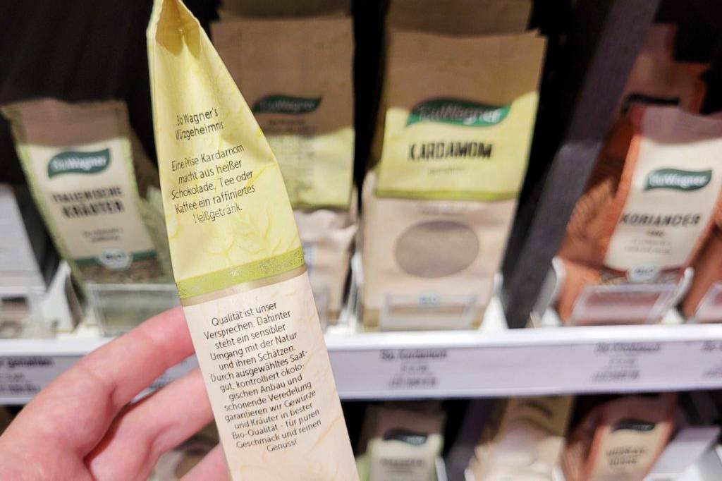 Kardamom getestet: Kardamom-Packung wird vor einem Regal auf der Seite in der Hand gehalten.