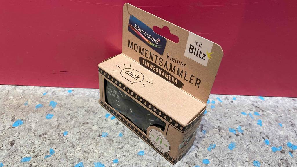 Einwegkamera im Test: Einwegkamera steht verpackt vor einer roten Wand.