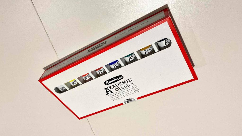 Ölfarben im Test: eine weiße Packung mit roter Umrandung Ölfarben in verschiedenen Farben liegend auf weißer Fläche.