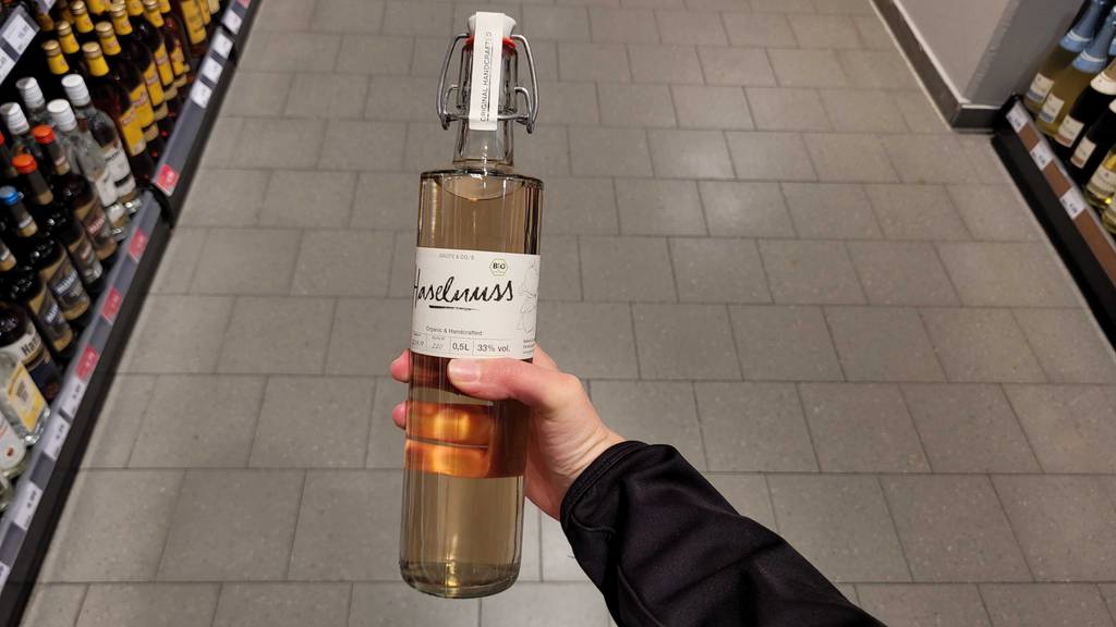 Eine Hand hält den getesteten Nusslikör in einer transparenten Flasche vor einem Verkaufsregal.