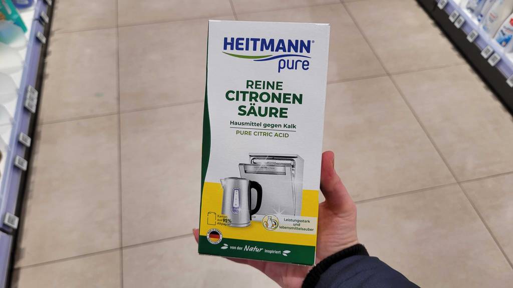 Eine Hand hält eine Packung mit getesteter Zitronensäure über dem Boden in einem Drogeriemarkt.