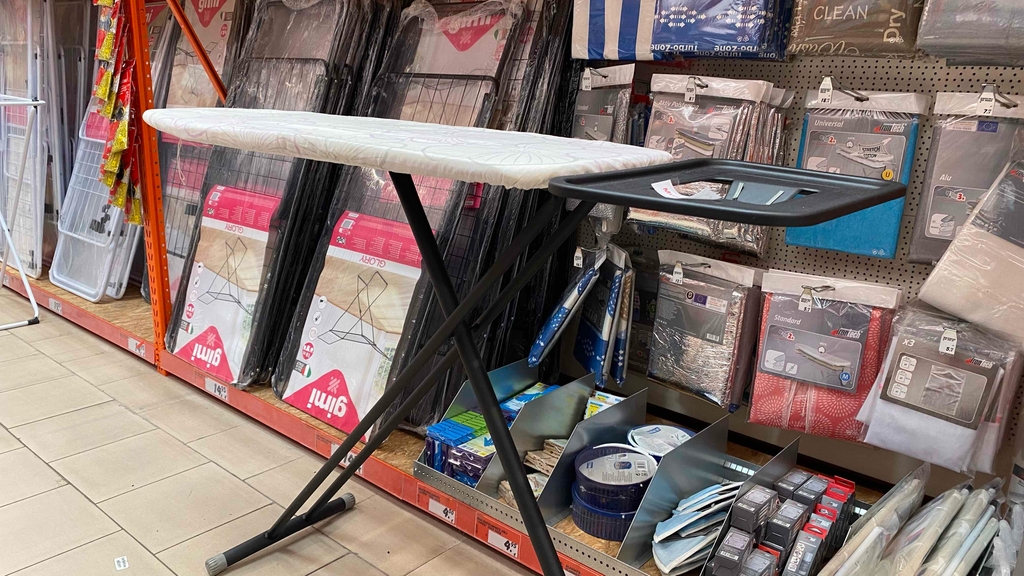 Bügelbrett mit weißem Bezug und schwarzem Gestell steht vor einem Regal.