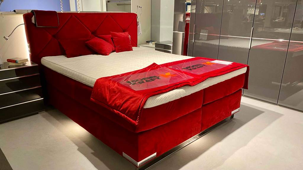 Boxspringbett-160x200 getestet: gesamtes Boxspringbett mit rotem Bettrahmen und roten Kissen darauf in einem Geschäft ausgestellt.