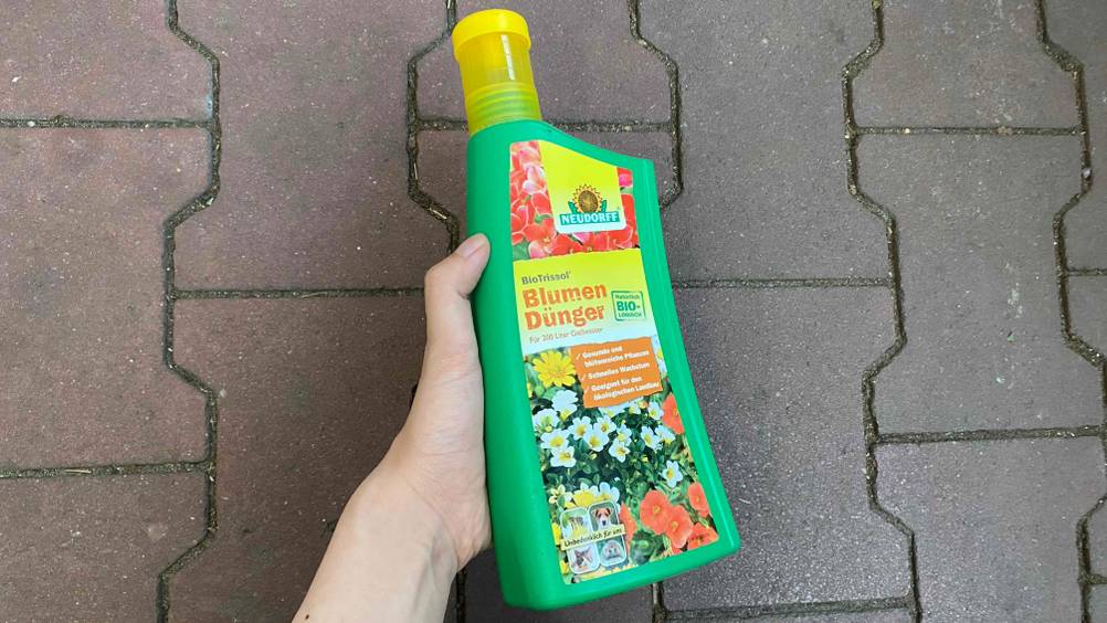 Blumendünger im Test: Eine Hand hält eine Flasche Dünger vor eine graue Fläche