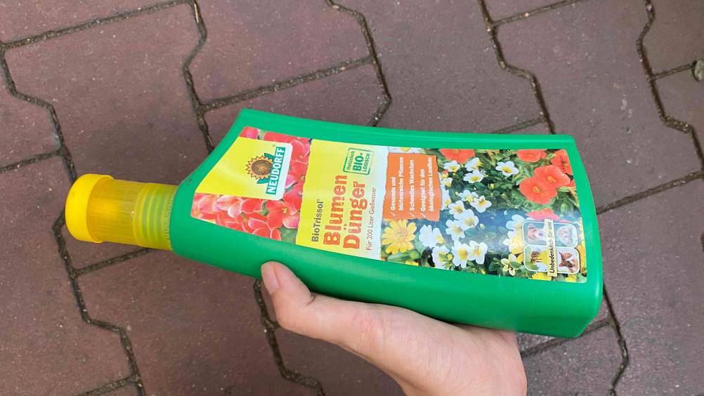 Blumendünger im Test: Eine Hand hält eine Flasche horizontal Dünger vor eine graue Fläche