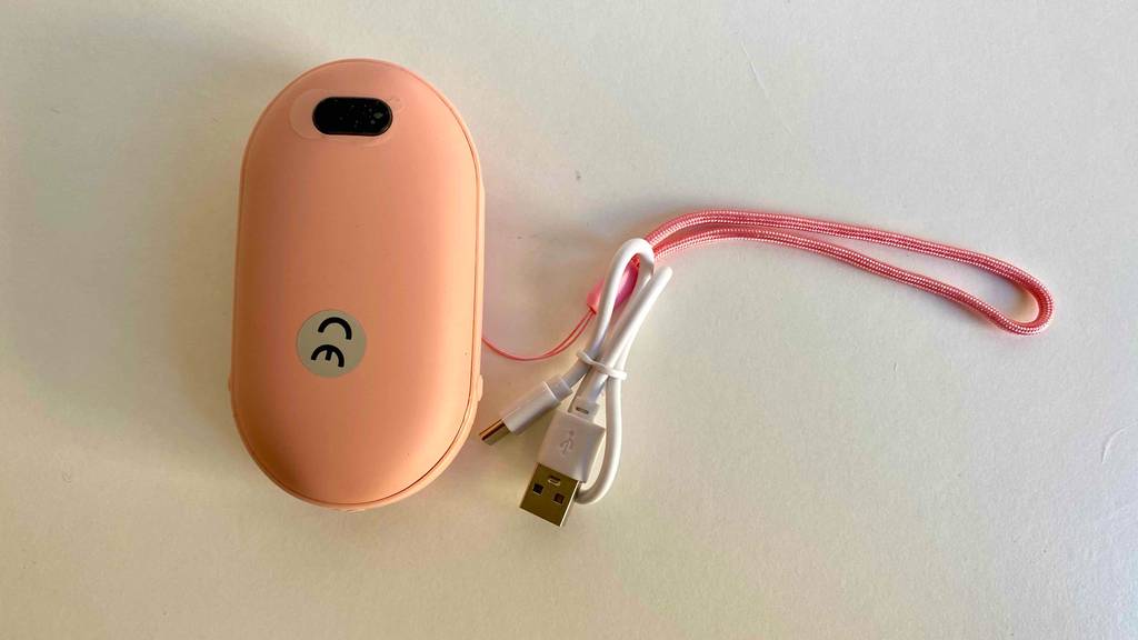 Ein getesteter USB-Handwärmer mit einem Band in Rosa liegt auf einer weißen Fläche neben dem Netzstecker.