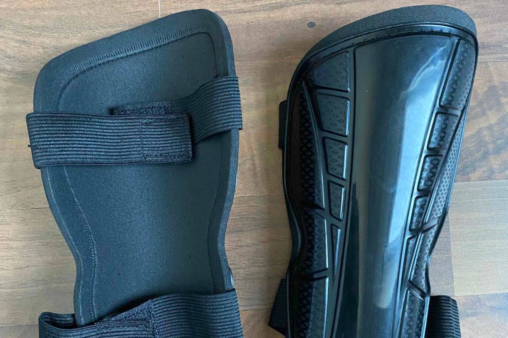 Zwei schwarze getestete Schienbeinschoner auf Holzboden, mit Sicht auf äußere Hartschalung und gepolsterter Rückseite inklusive elastischem Klettverschluss.